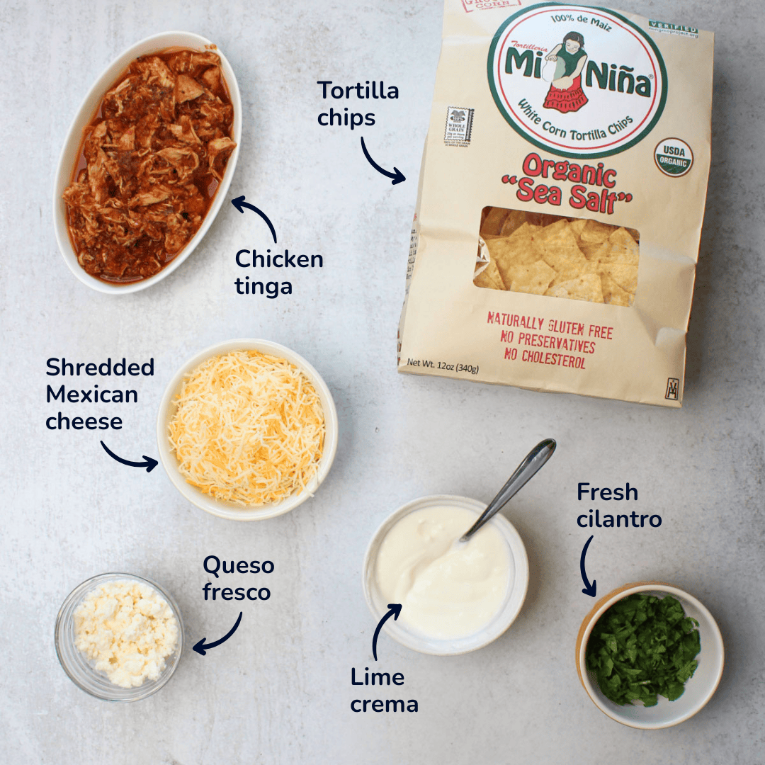 The ingredients needed to make chicken tinga nachos: chicken tinga, tortilla chips, shredded mexican cheese, queso fresco, lime crema, and fresh cilantro.