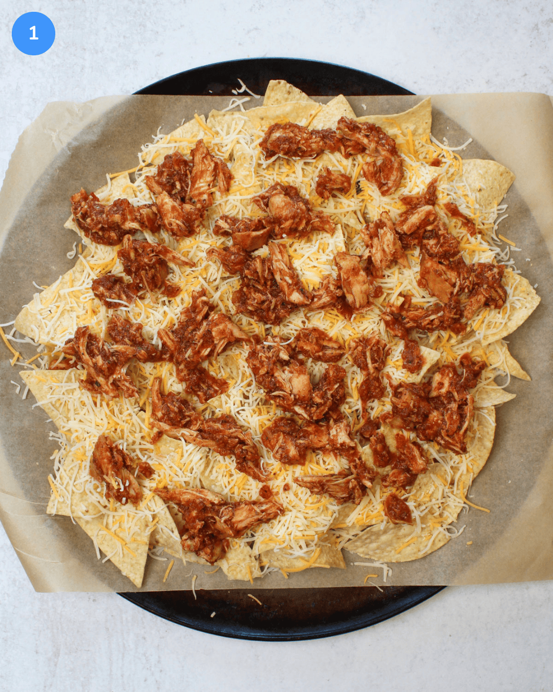 How to make chicken tinga nachos step 1: chips on a sheet pan topped with mexican cheese and chicken tinga.
