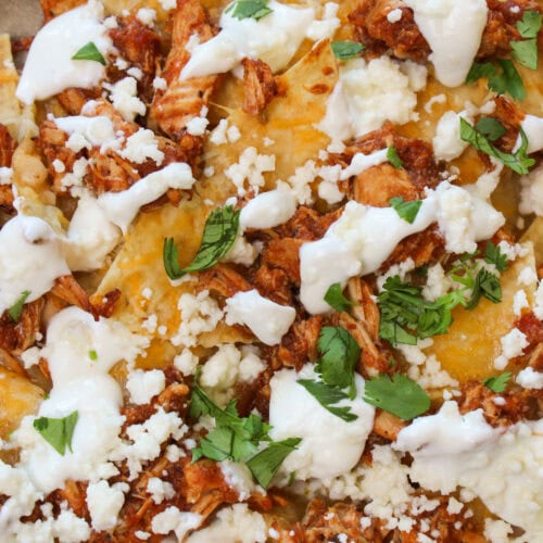 Chicken tinga nachos made with shredded mexican cheese, crockpot chicken tinga, queso fresco, lime crema, and cilantro.