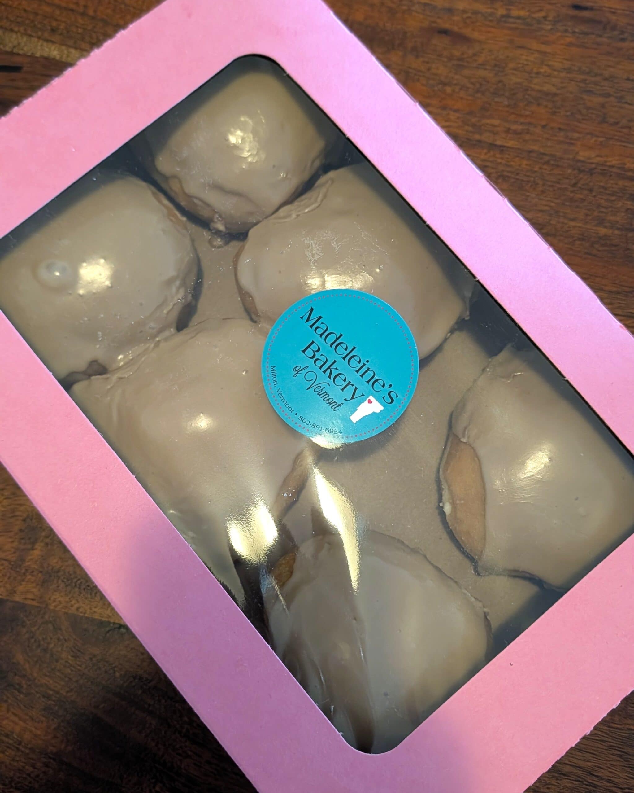 A pink box with a transparent lid full of 6 freshly made maple frosted donuts.