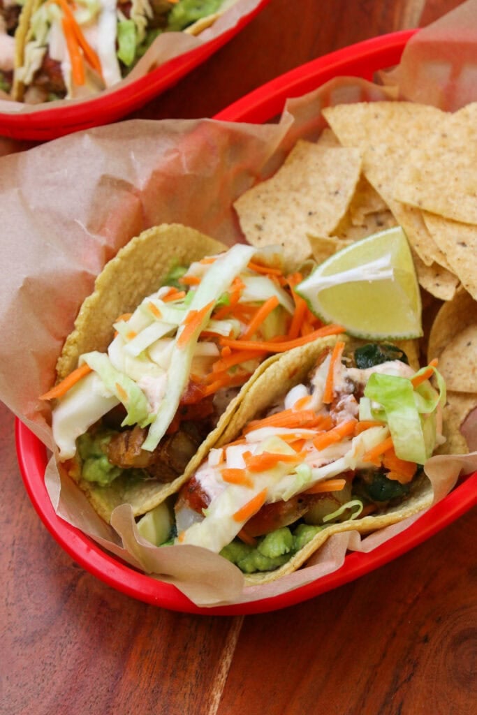 A red takeout basket lined with parchment serving two freshly made potato poblano tacos and tortilla chips.