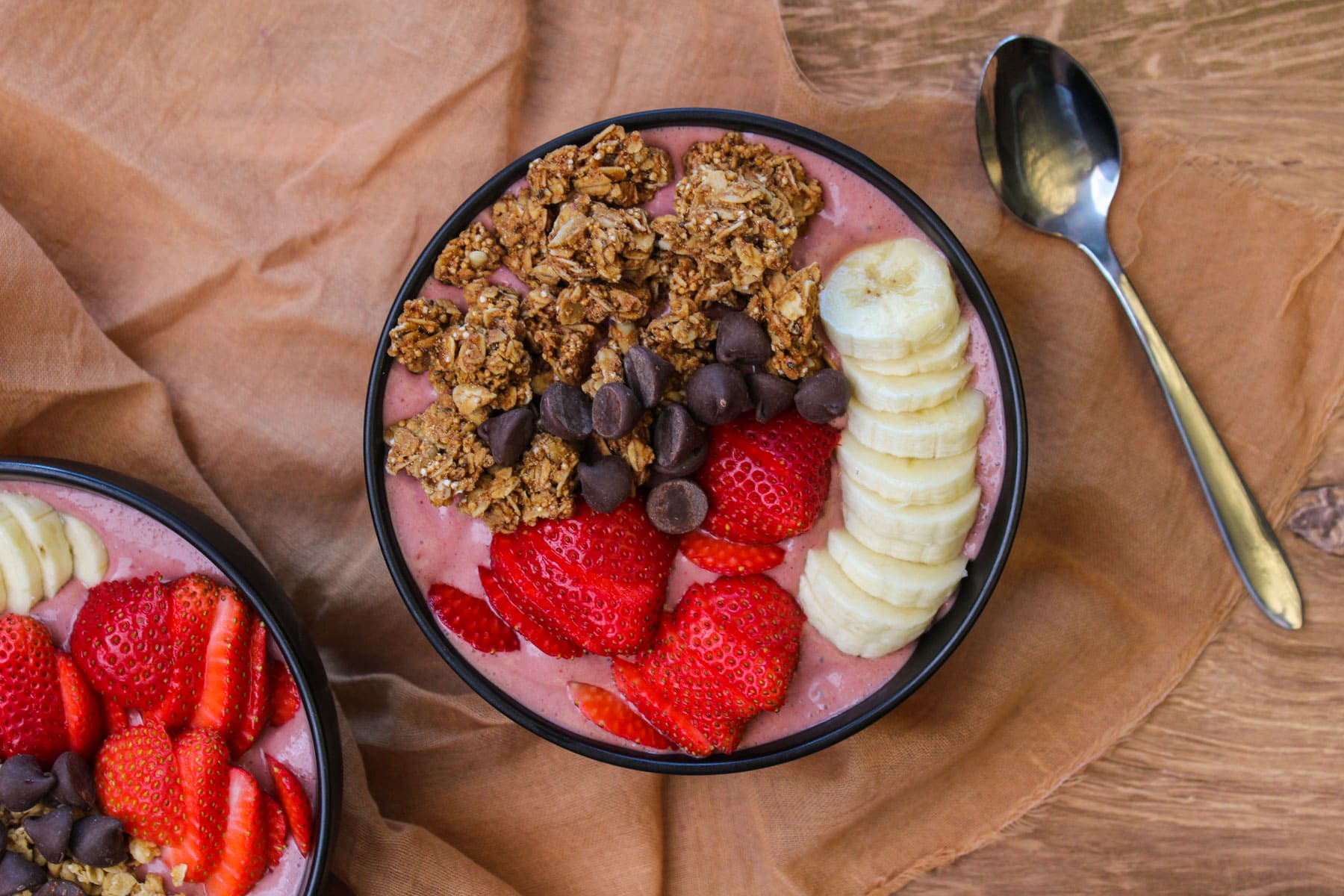 An acai smoothie bowl topped with fresh strawberries, banana slices, dark chocolate chips, and granola served in a black stone bowl.