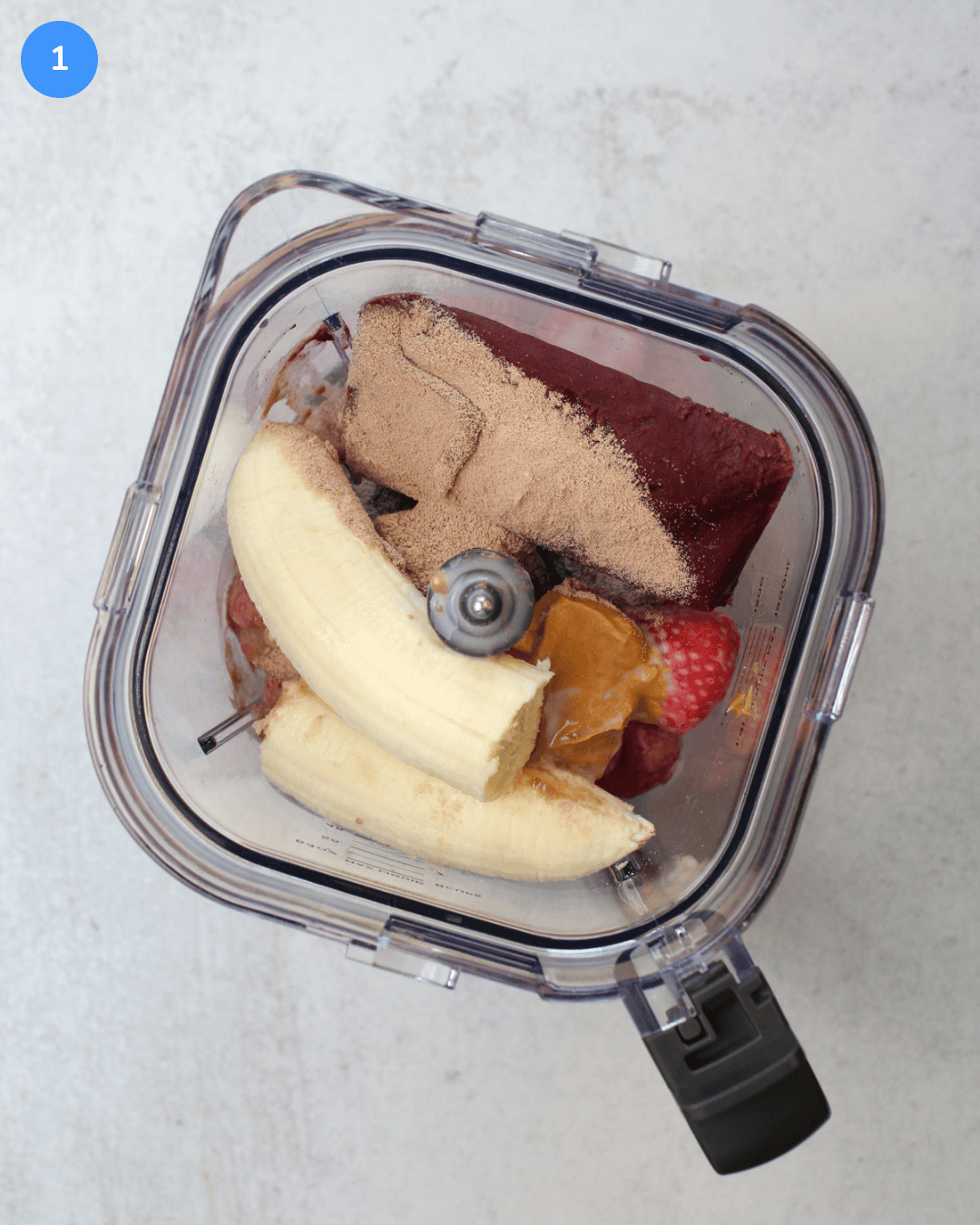 A blender filled with frozen acai, chocolate protein powder, peanut butter, strawberries, banana, and almond milk.
