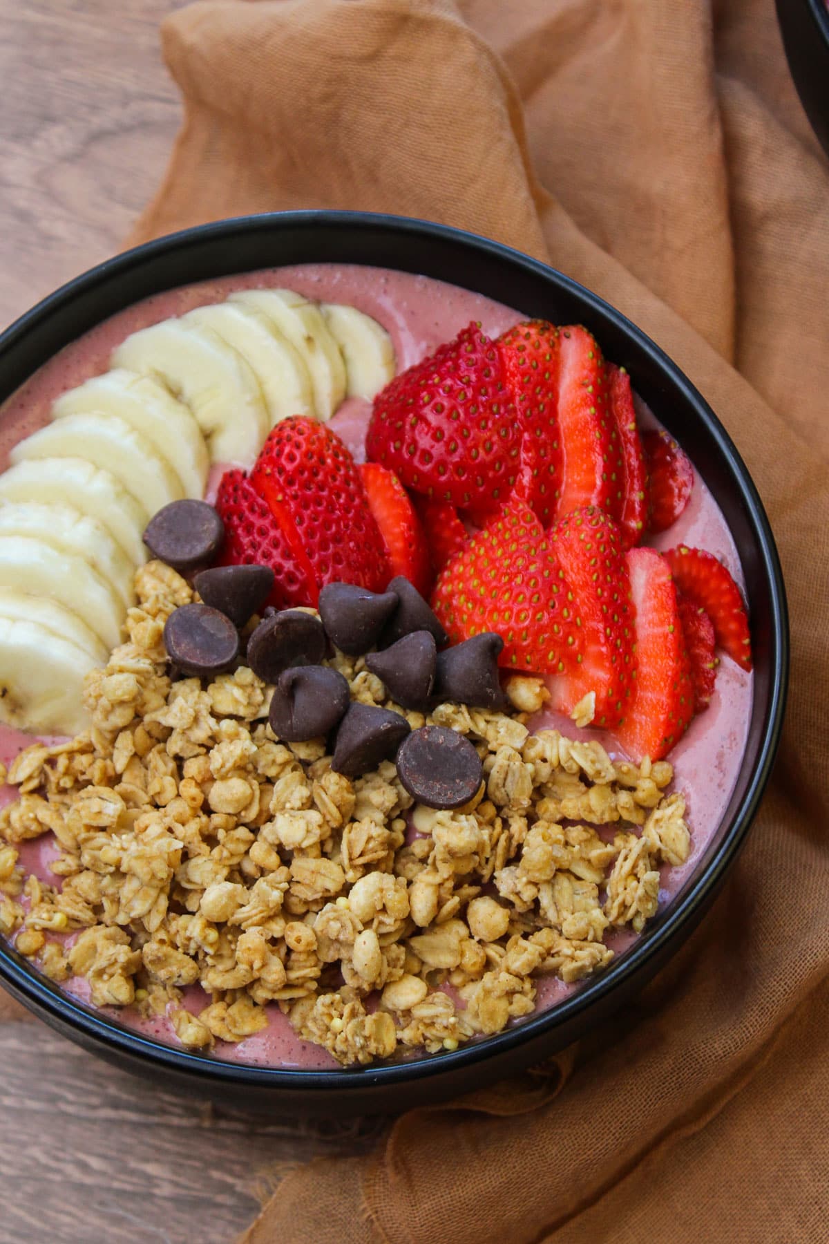 A freshly made acai smoothie bowl topped with fresh fruit, granola, and dark chocolate chips in a black bowl.