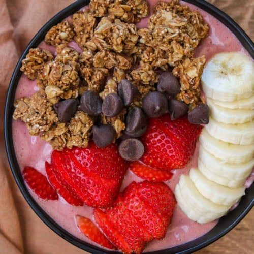A pink acai smoothie bowl with granola, chocolate chips, and fresh strawberry and banana slices on top.