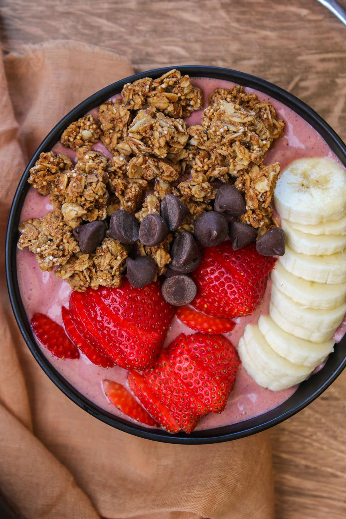 A pink acai smoothie bowl with granola, chocolate chips, and fresh strawberry and banana slices on top.