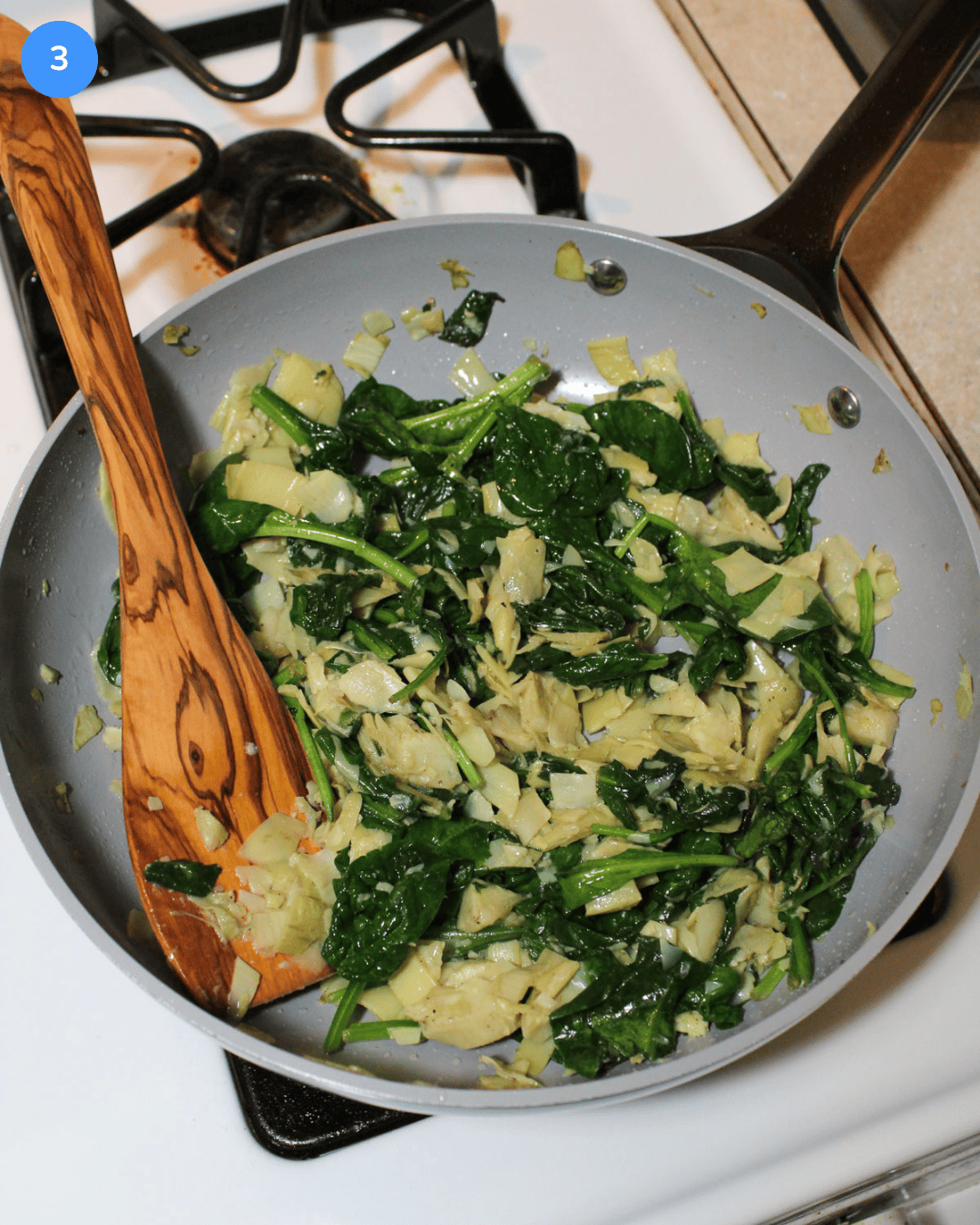Artichokes and spinach cooking in a ceramic frying pan on the stove.