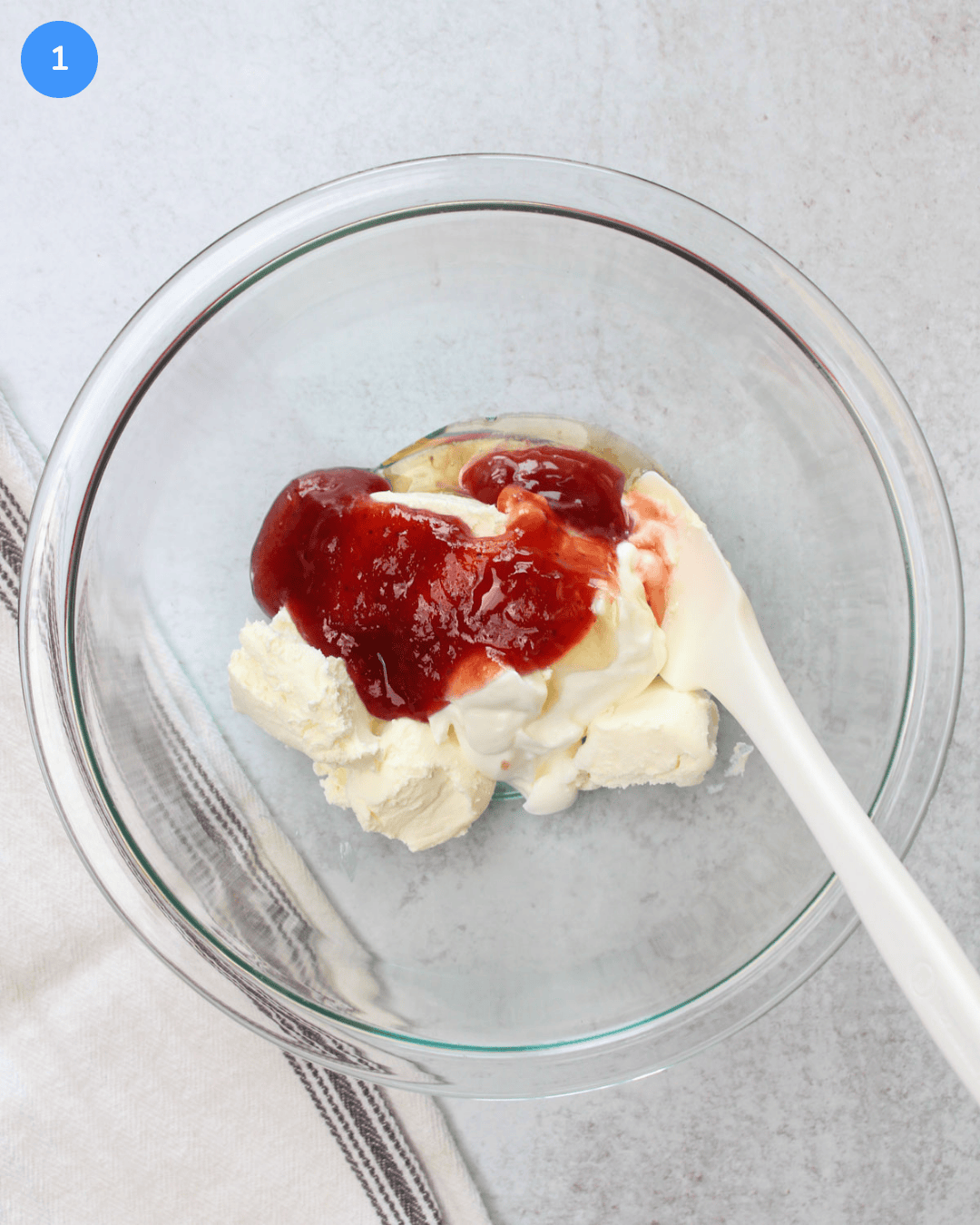 A glass mixing bowl filled with cream cheese, greek yogurt, strawberry jam, and honey.