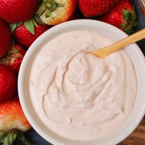 A white ceramic bowl of strawberry fruit dip with a wooden serving spoon surrounded by ripe strawberries in a blue stoneware bowl.