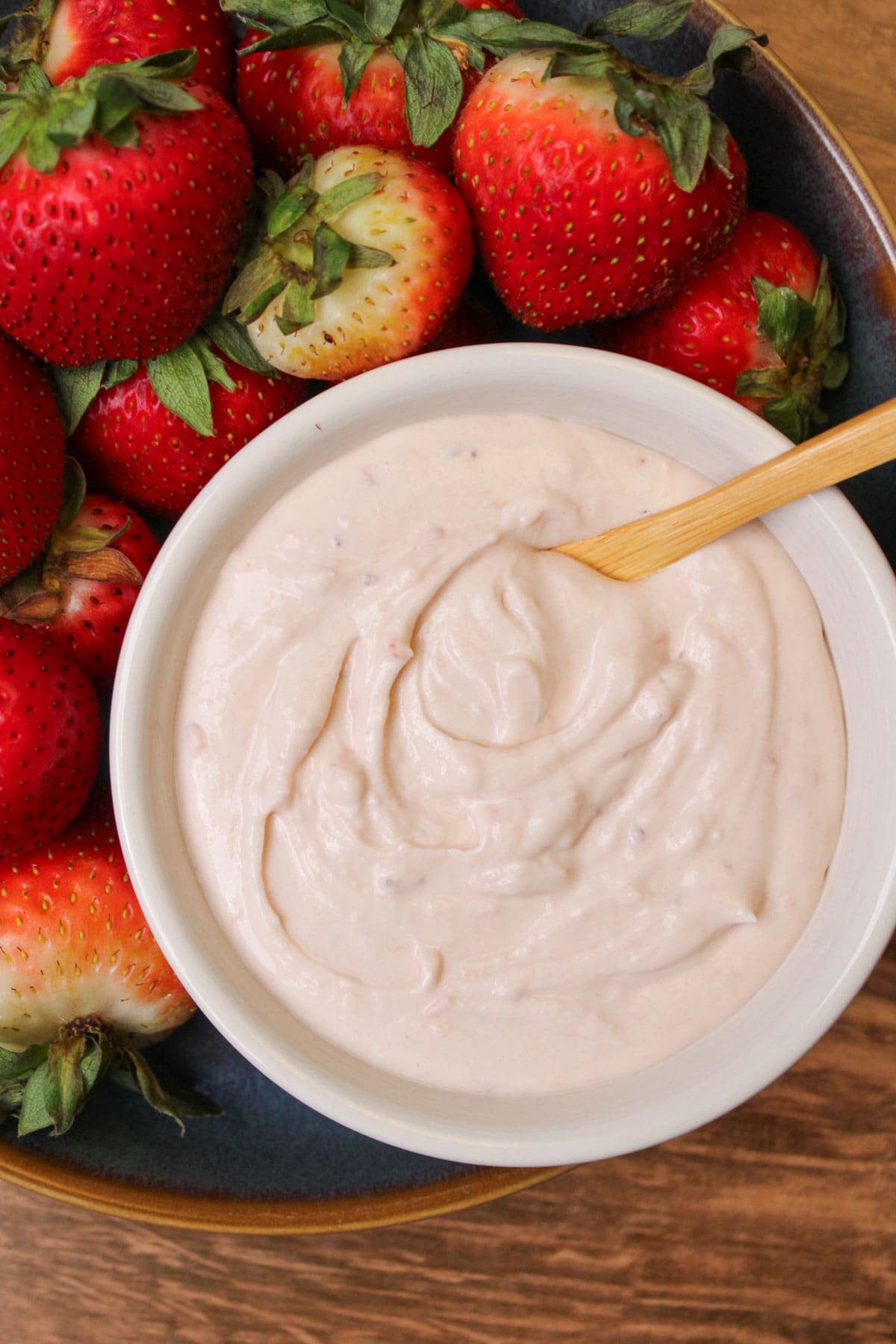 A white ceramic bowl of strawberry fruit dip with a wooden serving spoon surrounded by ripe strawberries in a blue stoneware bowl.