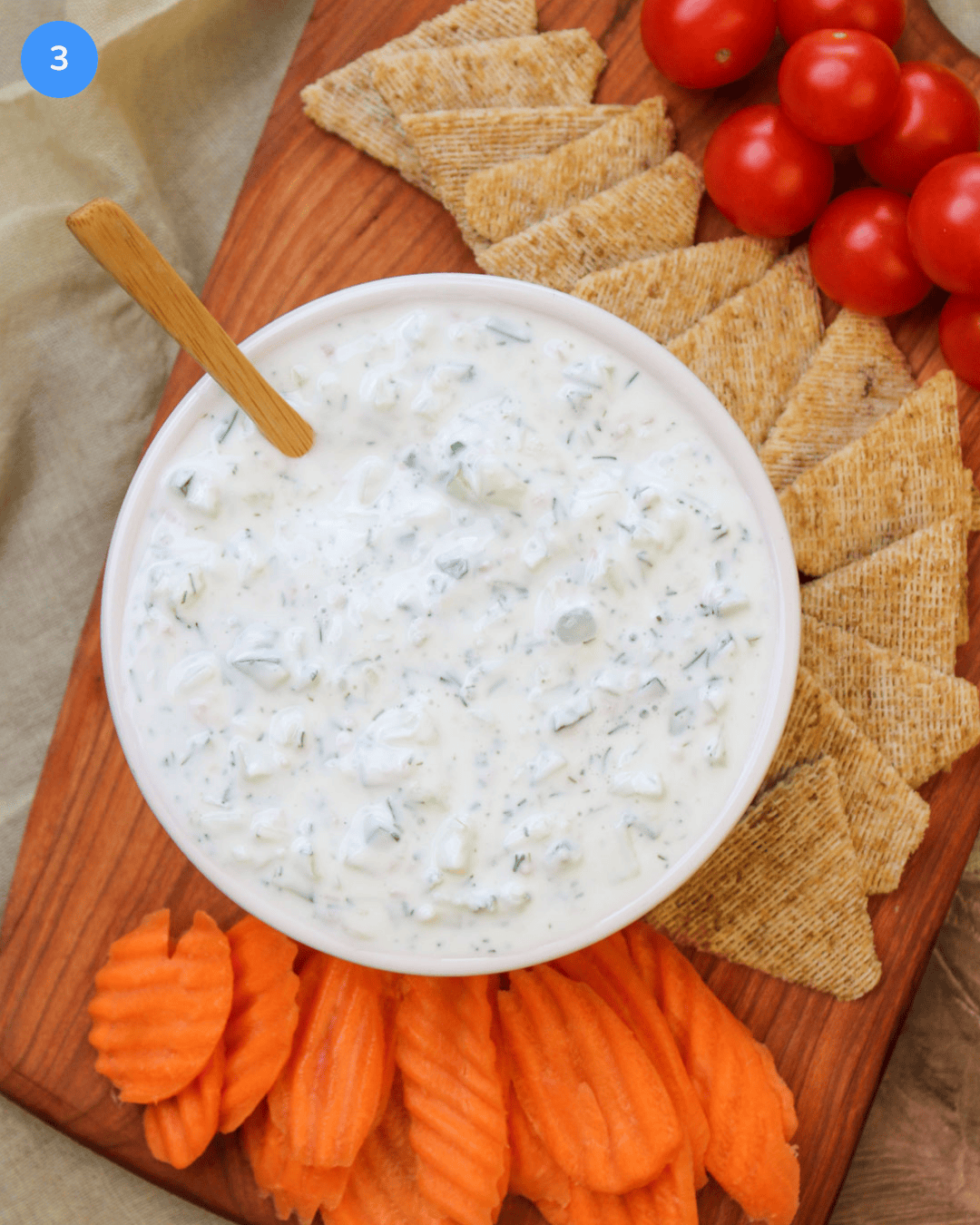 A white ceramic bowl of freshly made tzatziki with a wooden serving spoon on a wooden board surrounded by cherry tomatoes, carrot chips, and triscuit thins.