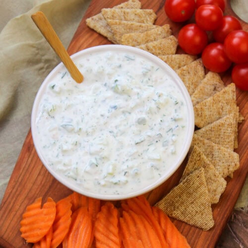 A serving platter of freshly made tzatziki, cherry tomatoes, wheat crackers, and carrot chips.