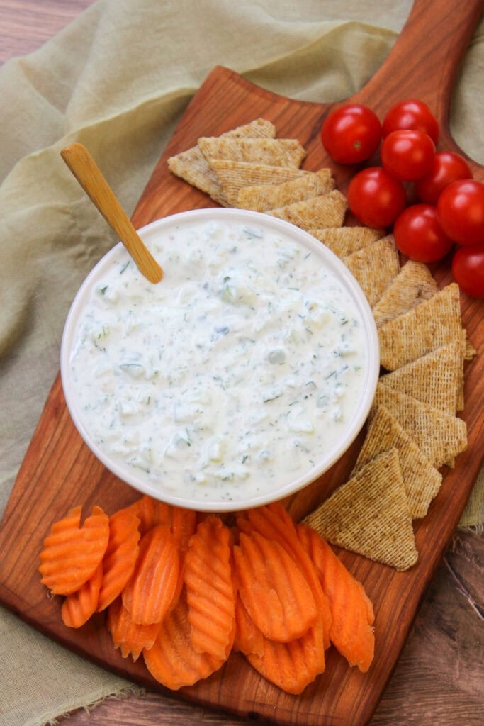 A serving platter of freshly made tzatziki, cherry tomatoes, wheat crackers, and carrot chips.