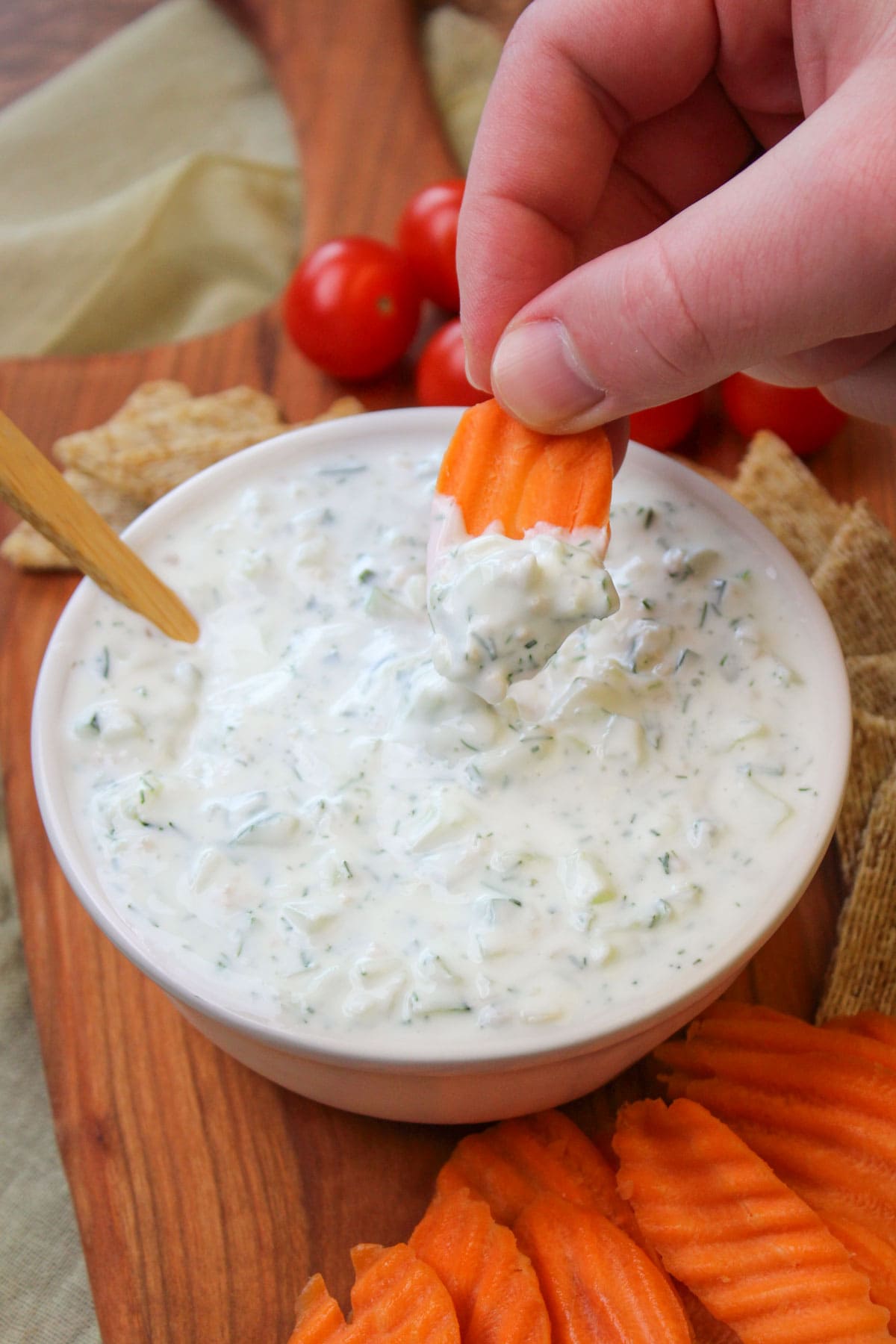 A carrot chip being dipped into a white ceramic bowl of freshly made tzatziki with dill.
