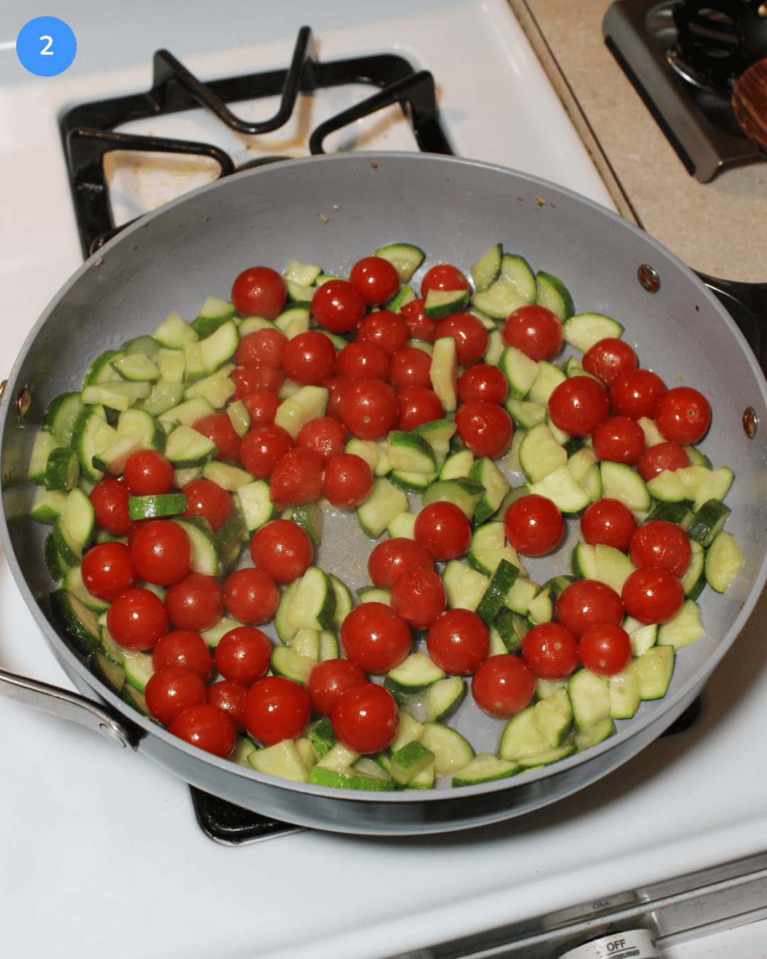 A ceramic saute pan with cherry tomatoes and quartered sliced zucchini cooking in olive oil and garlic.