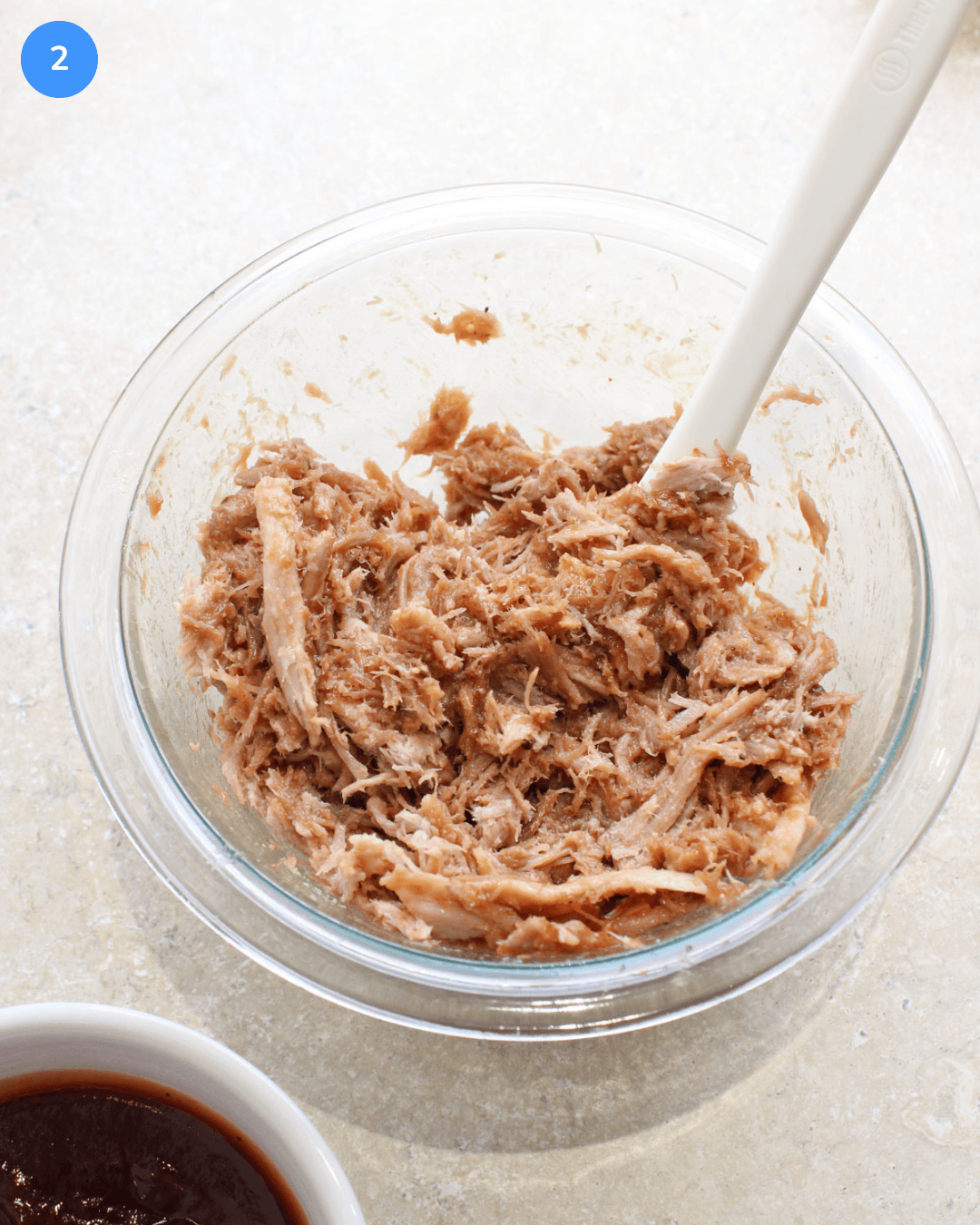 A glass mixing bowl of pulled pork and barbecue sauce being stirred together.