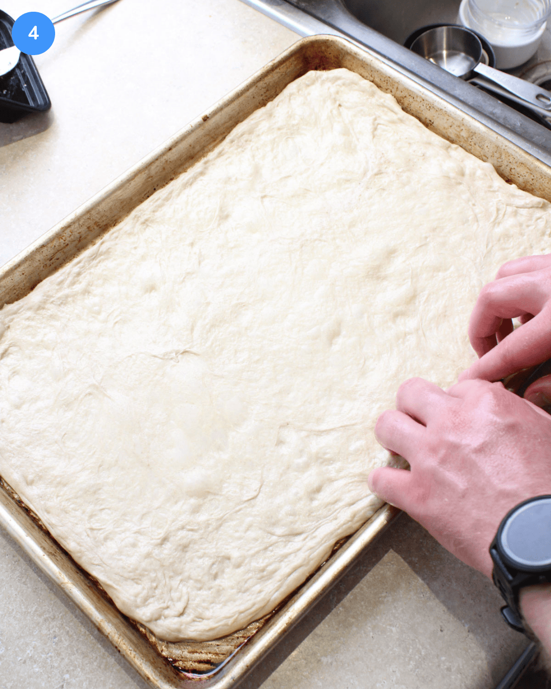 Pizza dough being stretched into a rectangle on an aluminum sheet pan.