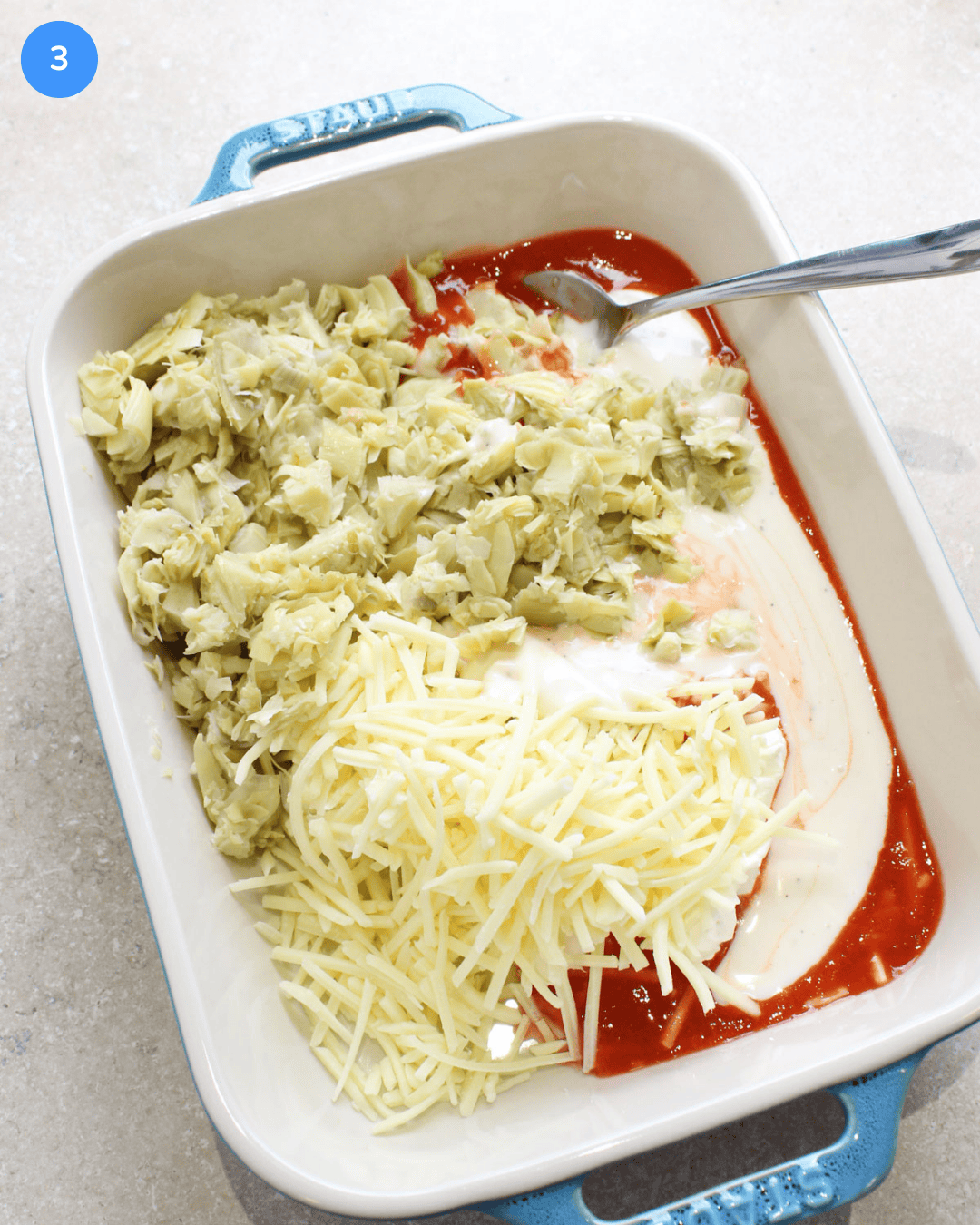 A ceramic baking dish with cream cheese, shredded cheddar, chopped artichokes, ranch dressing, and hot sauce in it.