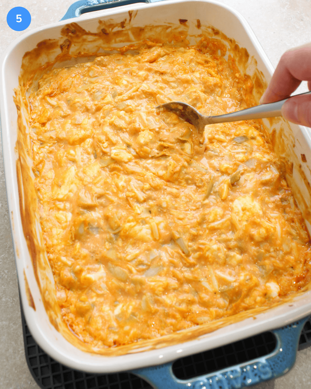 A ceramic baking dish of hot, cheesy buffalo artichoke dip being stirred.