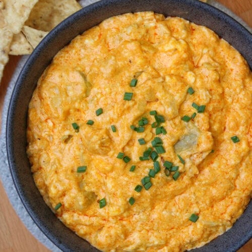A blue stone bowl of freshly baked buffalo artichoke dip, served with tortilla chips on a wooden platter.