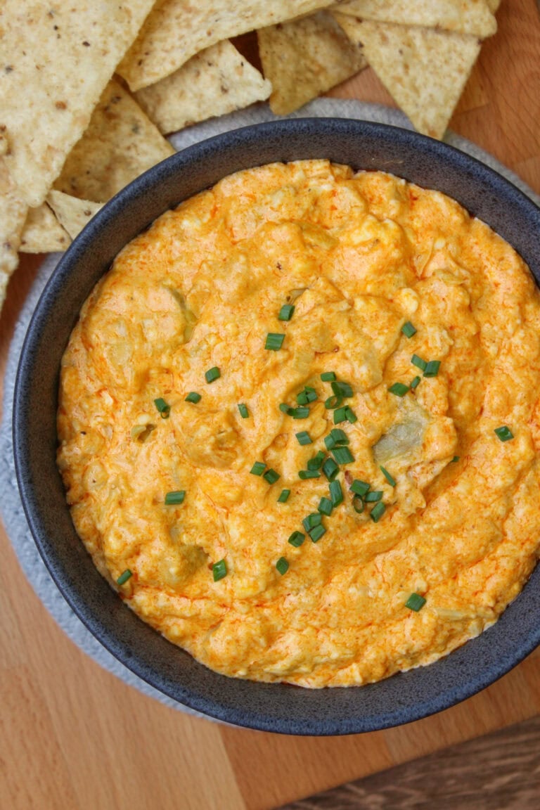 A blue stone bowl of freshly baked buffalo artichoke dip, served with tortilla chips on a wooden platter.