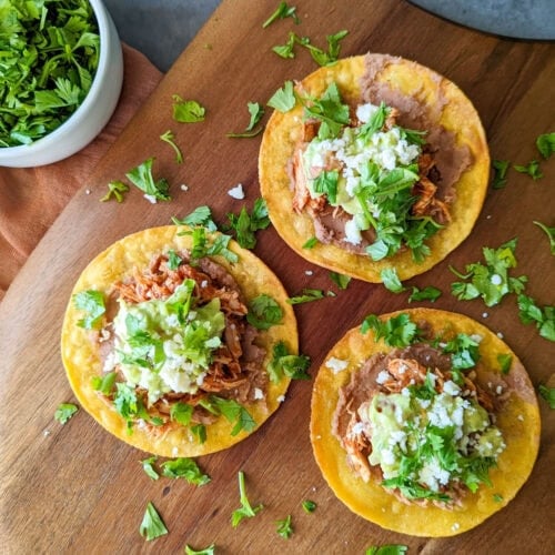 A wood platter of three chicken tinga tostadas with refried beans, guacamole, queso fresco, and fresh cilantro.