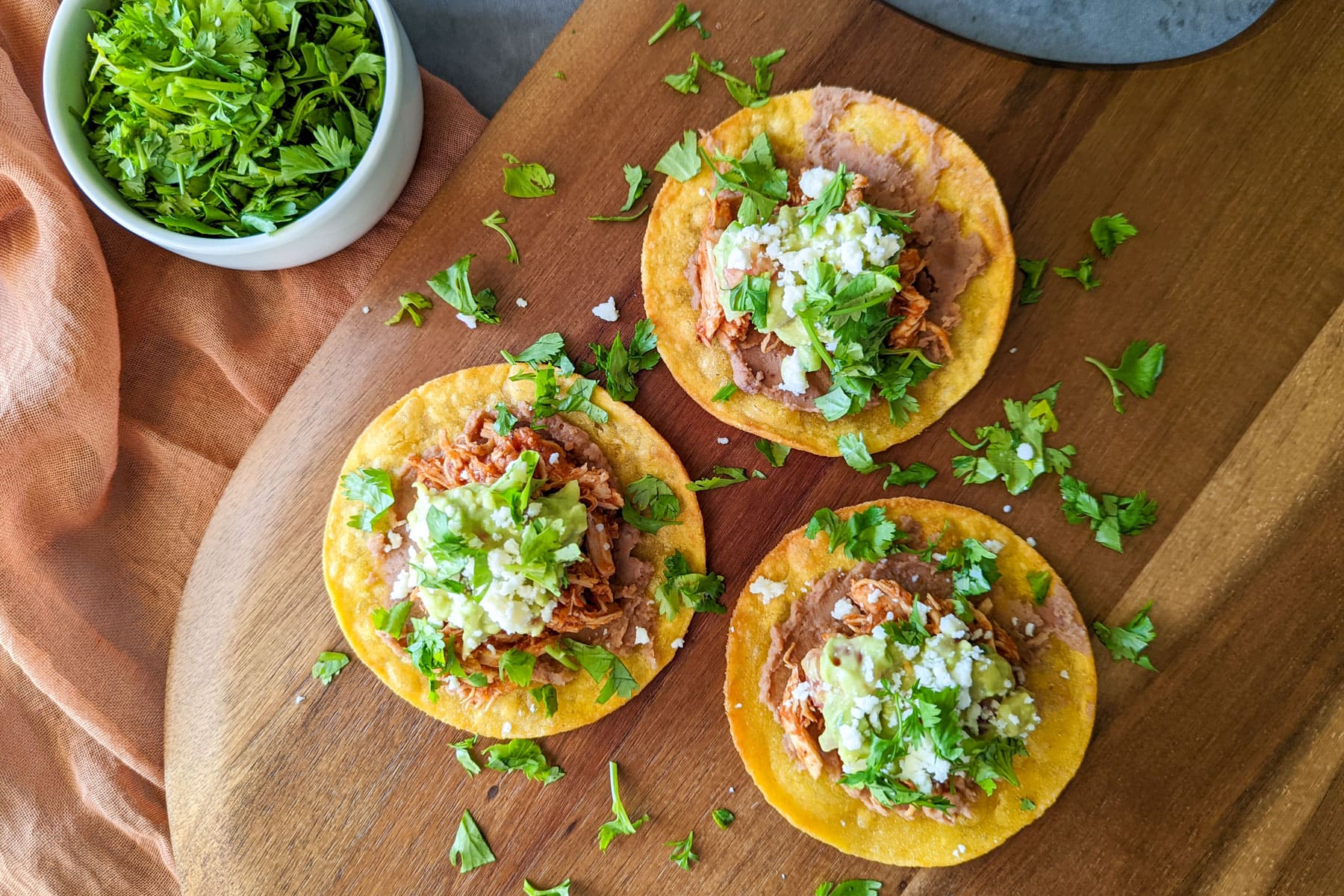 A wood platter of three chicken tinga tostadas with refried beans, guacamole, queso fresco, and fresh cilantro.