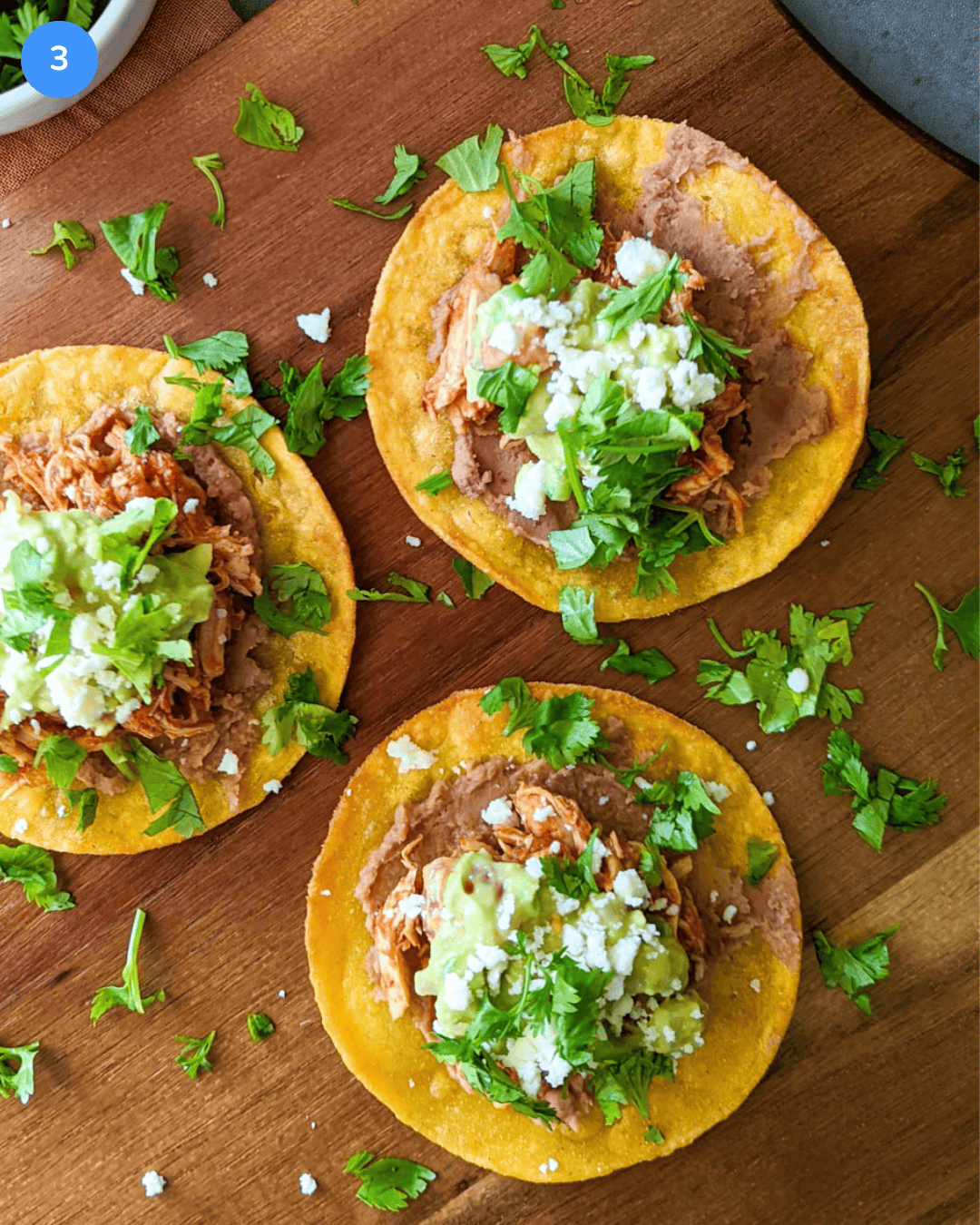 Three freshly assembled chicken tinga tostadas with mashed avocado, refried beans, queso fresco, and cilantro on a wood serving platter.