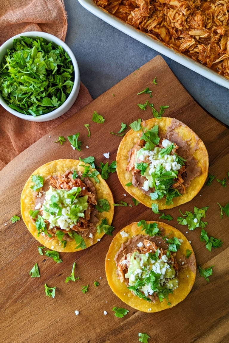 A wood platter of three freshly made chicken tinga tostadas, served alongside fresh cilantro and a dish of chicken tinga.