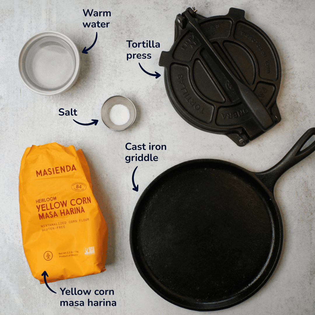 The ingredients and equipment needed to make homemade corn tortillas: masa harina, water, salt, a tortilla press, and a cast iron griddle.