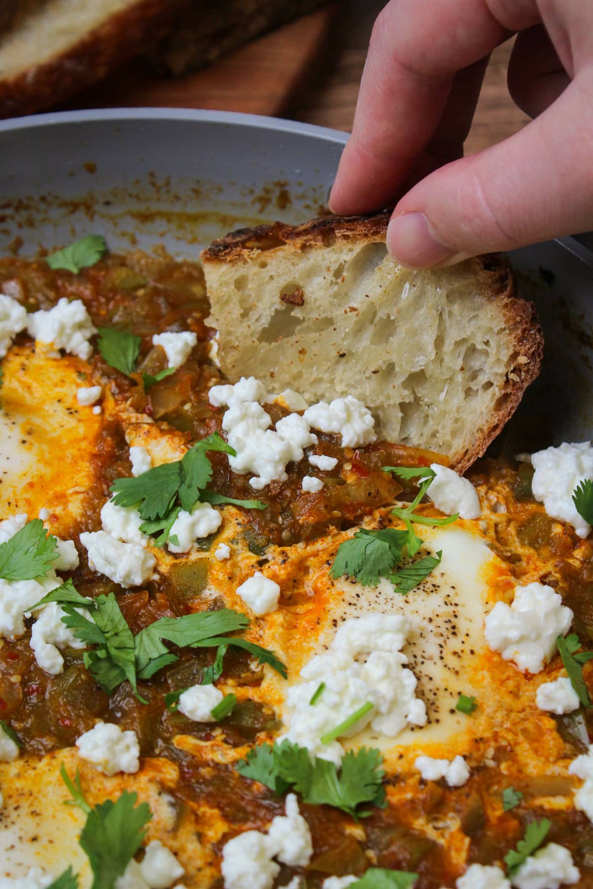 A slice of crusty white bread being dipped into a skillet of freshly made green shakshuka with cilantro and goat cheese.