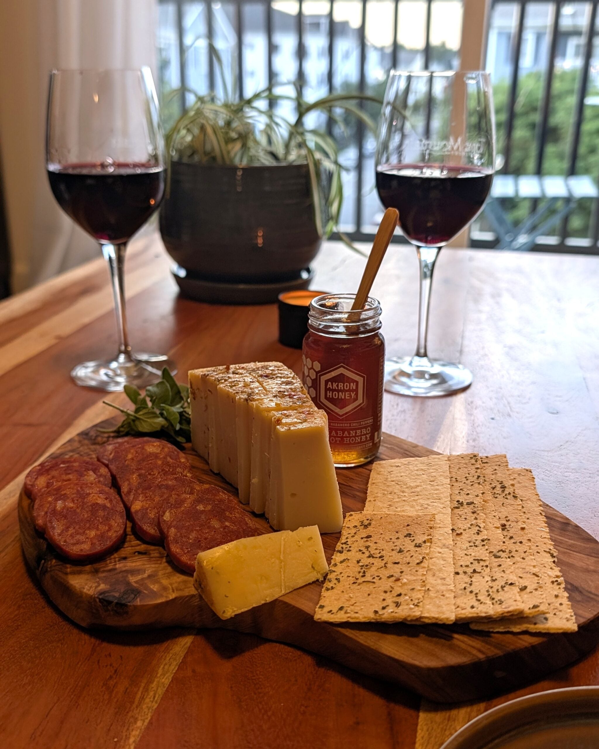Two glasses of red wine and a cheeseboard with pepperoni, crackers, and hot honey on a wooden coffee table.