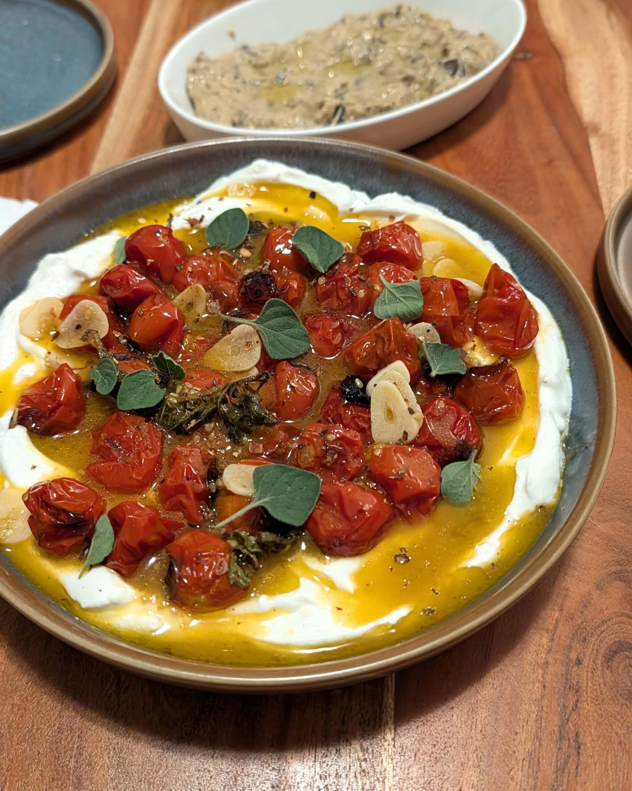 A blue stone bowl filled with greek yogurt and roasted tomatoes with garlic and fresh oregano on a wood table.
