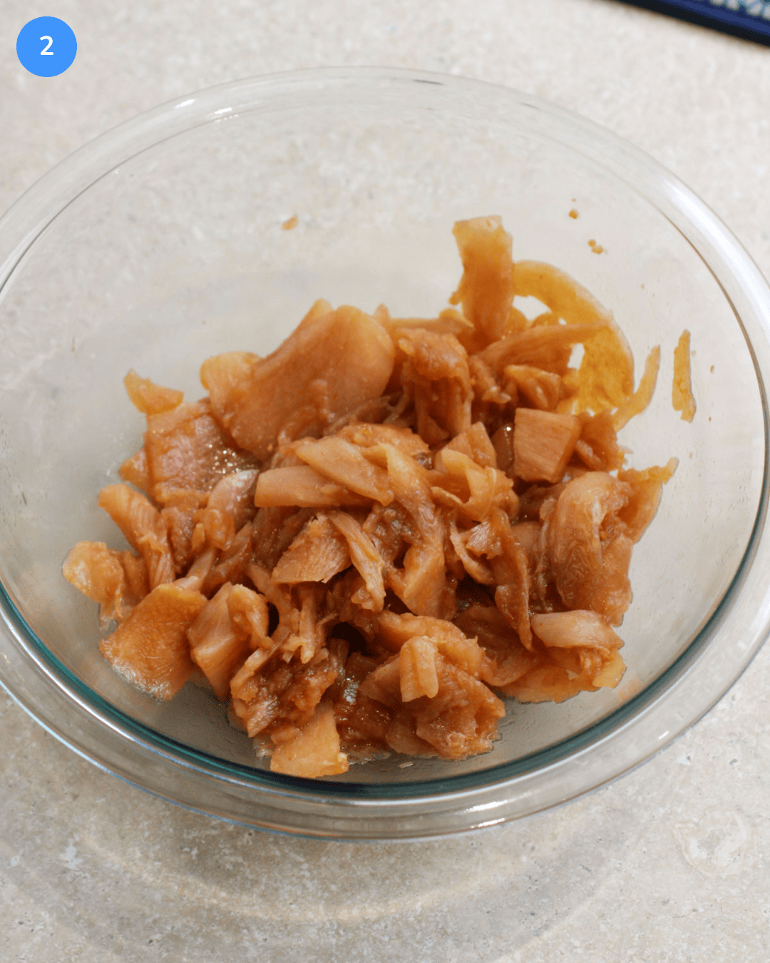A glass mixing bowl of thinly shaved chicken marinating in a soy sauce mixture.