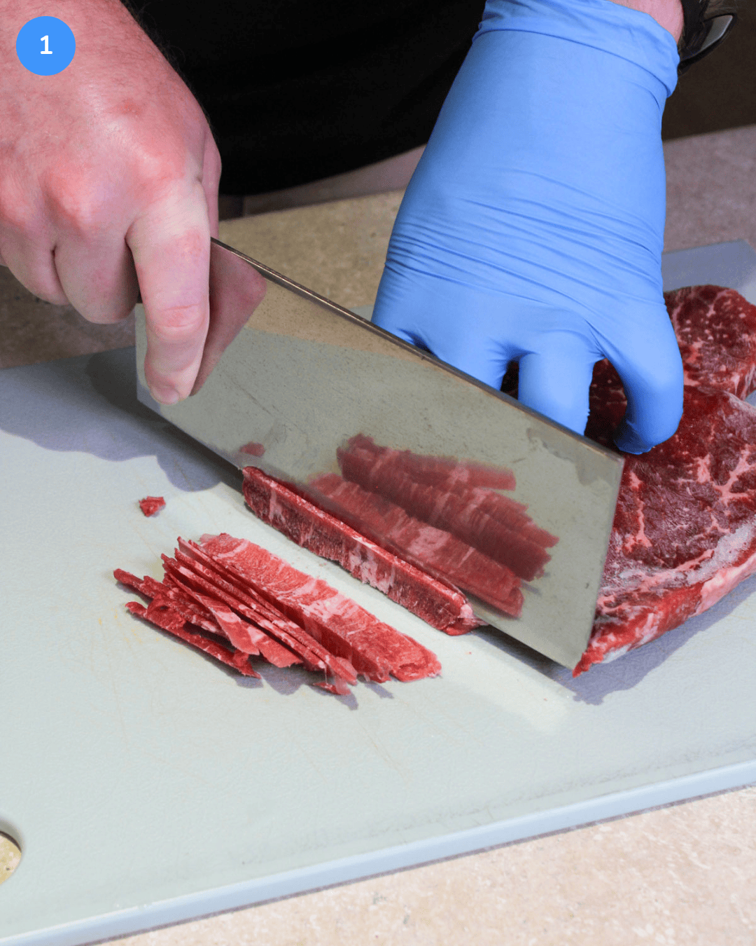 Raw, partially frozen flank steak being sliced thin with a large chinese chef's knife.