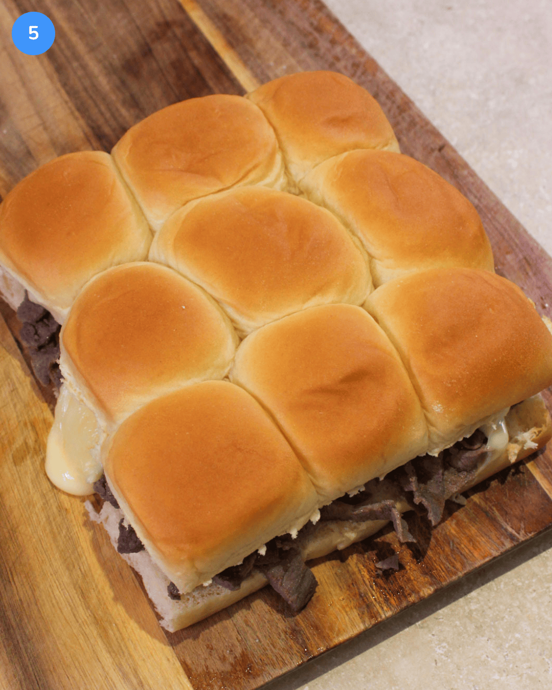 A pack of 9 pull-apart philly cheesesteak sliders on a wood cutting board.