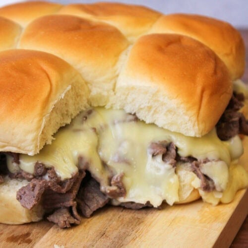 A wood serving board of gooey, cheesy philly cheesesteak sliders on hawaiian rolls.