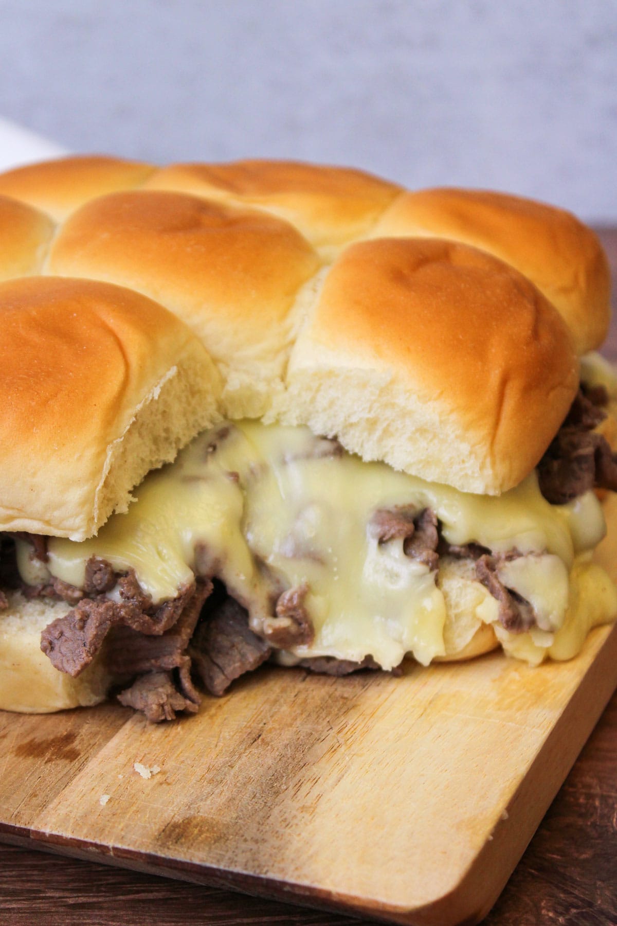 A wood serving board of gooey, cheesy philly cheesesteak sliders on hawaiian rolls.