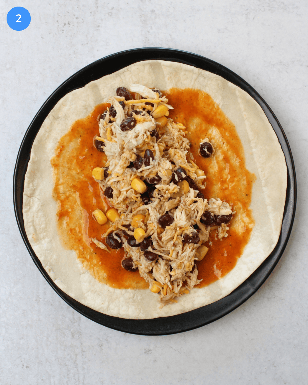 A flour tortilla on a black plate topped with red enchilada sauce and a black bean, corn, chicken, and cheese filling.