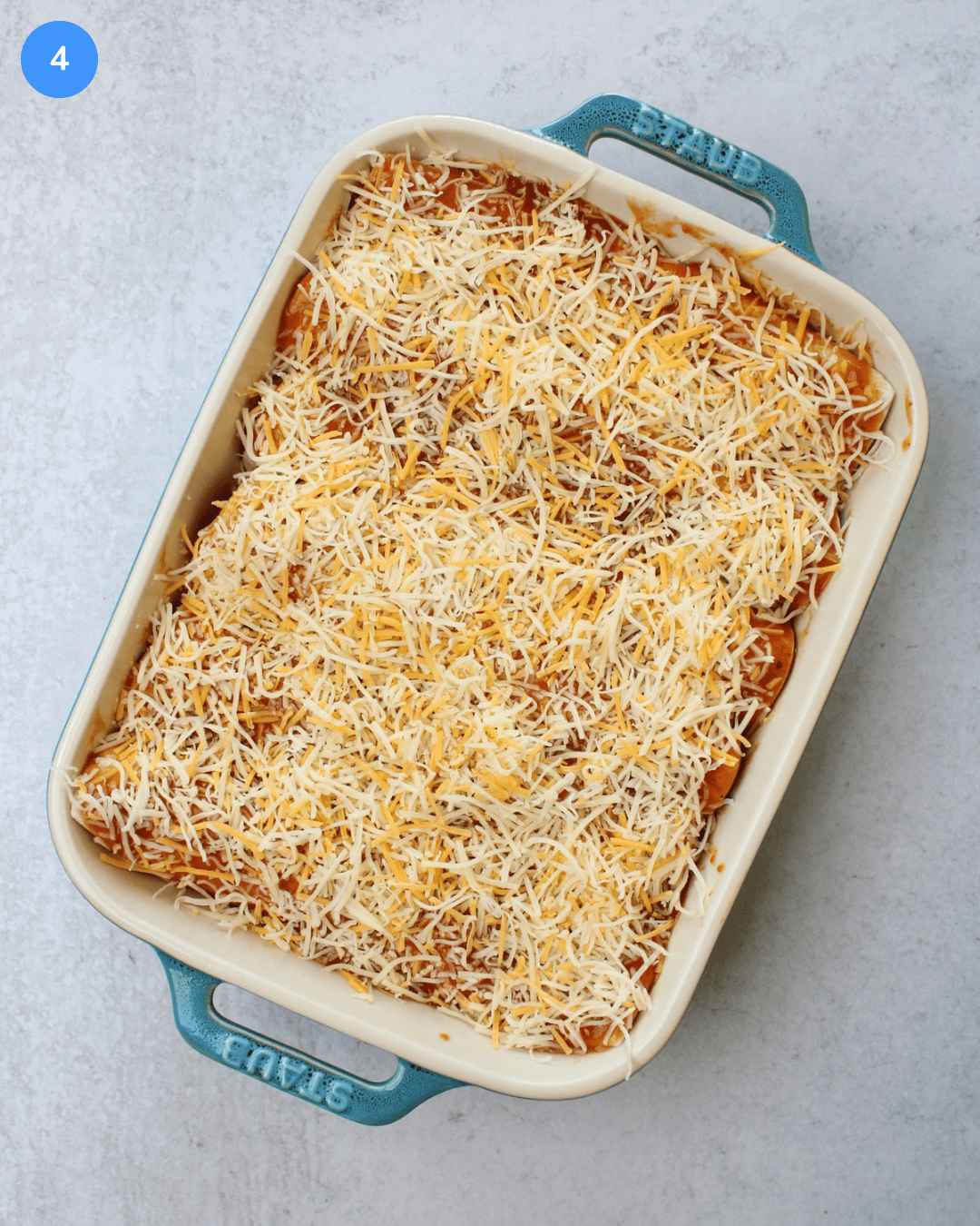 A ceramic baking dish of uncooked enchiladas topped with red sauce and shredded mexican cheese.