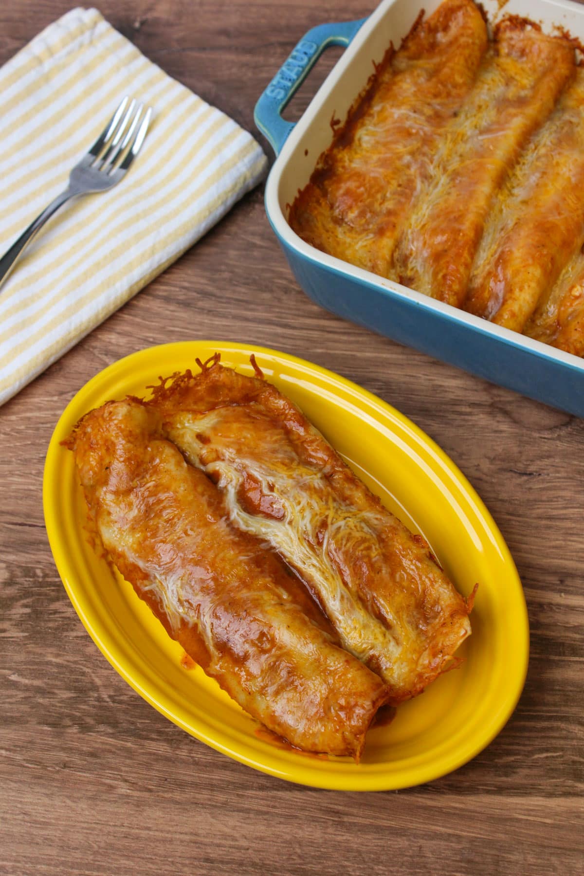 Two red chicken enchiladas on a yellow ceramic plate next to a blue ceramic baking dish of enchiladas.