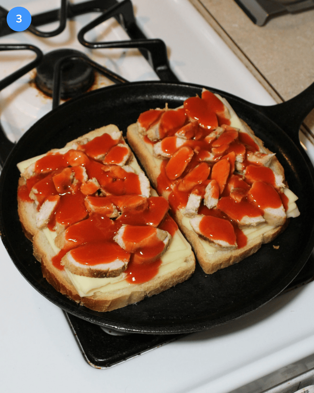 Two slices of sourdough topped with american cheese, grilled chicken, and frank's red hot on a cast iron griddle on the stove.